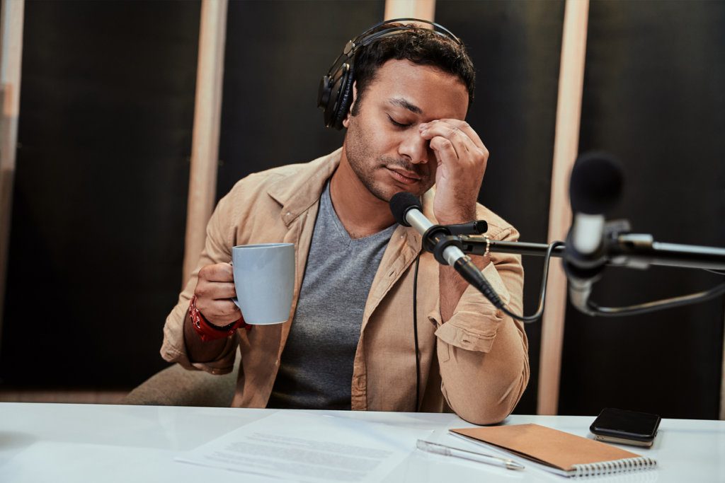 Portrait of young male radio host in headphones looking tired, exhausted, drinking coffee while getting ready for broadcasting in studio. Horizontal shot