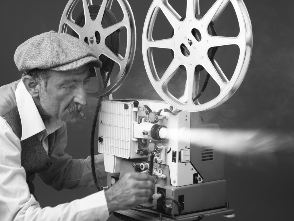 Senior man wearing shirt,waist and flat cap starting old-fashioned film projector. He is placed on the left side of the frame. Smoke and light beams come out of the projector lens. The photo is black and white and has horizontal composition.