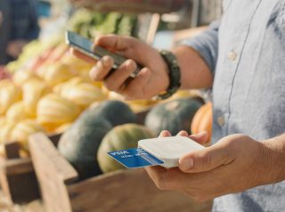 A vender holds the Square reader in one hand with a credit card inserted into it and his smartphone in the other. A vegetable market stand is in the background.