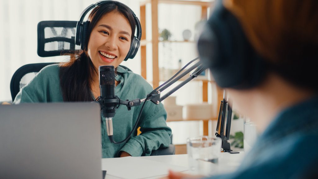 Woman radio host recording podcast using microphone wearing headphones interviewing guest sitting across from her.